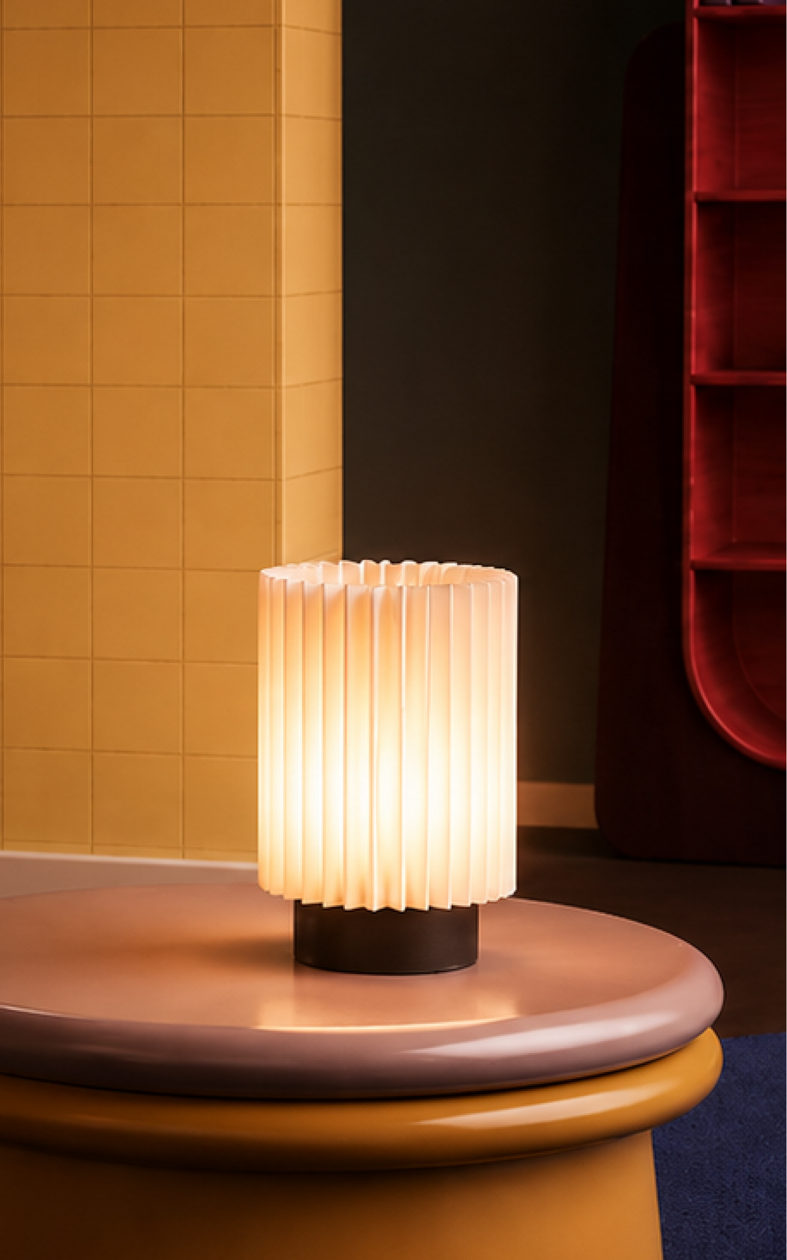 A modern interior scene with a low round coffee table in mustard yellow and taupe, topped with a glowing pleated white table lamp. To the left is a sculptural orange armchair with a pink cushion, set against a warm yellow tiled wall.